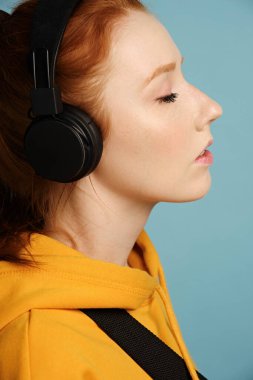 Kızıl saçlı, gözleri kulaklıkla kapalı ve sarı kapüşonlu bir kızın portresi..