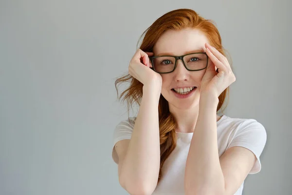 Una chica pelirroja con una camiseta blanca está de pie sobre un fondo gris sonriendo y sosteniendo las gafas . — Foto de Stock