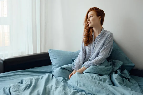 Çizgili pijamalı kızıl saçlı bir kız mavi bir yatakta oturur ve güneş ışığında pencereden dışarı bakar.. — Stok fotoğraf