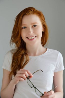 Beyaz tişörtlü güzel kızıl saçlı bir kız gri bir arka planda duruyor ve elinde gözlüklerle gülümsüyor.