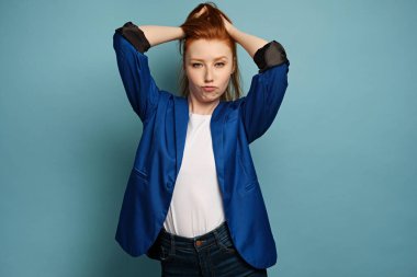 Mavi ceketli kızıl saçlı bir kız mavi arka planda duruyor, saçlarını topluyor ve kaşlarını kaldırıyor.