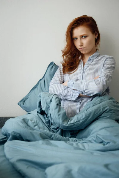 Pijamalı kızıl saçlı kız mavi bir yatakta oturuyor ve kareye hoşnutsuzca bakıyor, kollarını göğsüne koyuyor ve dudaklarını buruşturuyor. — Stok fotoğraf