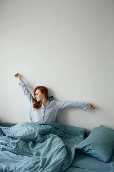 Pijamalı kızıl saçlı kız mavi bir yatakta oturur ellerini yana uzatarak beyaz duvarın arka planına yaslanır.. — Stok fotoğraf