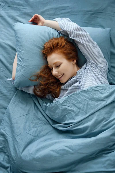 Çizgili pijamalı kızıl saçlı bir kız mavi bir yatakta uzanıyor. Çerçeve üstte. — Stok fotoğraf