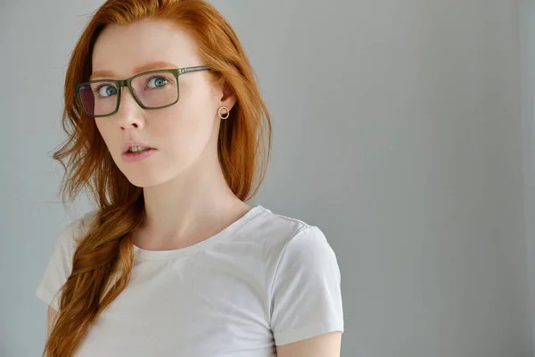 Ein rothaariges Mädchen mit Brille und weißem T-Shirt steht auf einem grauen Hintergrund in halber Drehung und blickt überrascht in den Rahmen. — Stockfoto