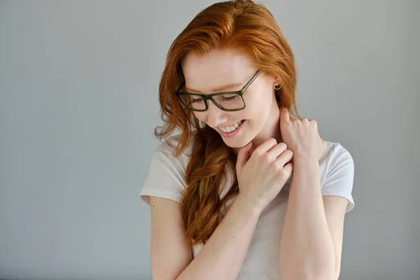 Una chica pelirroja con una camiseta blanca y gafas se para sobre un fondo gris y se ríe con la cabeza baja. . — Foto de Stock