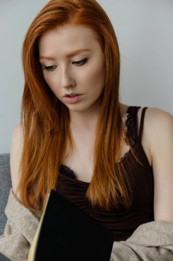 Koyu renk bluzlu güzel kızıl saçlı kız siyah bir kitap okuyor, yüzüne odaklanıyor..