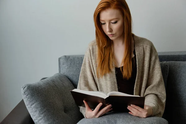 Red-haired girl in a warm cape sits on the sofa and carefully reads the black book, lowering her head.