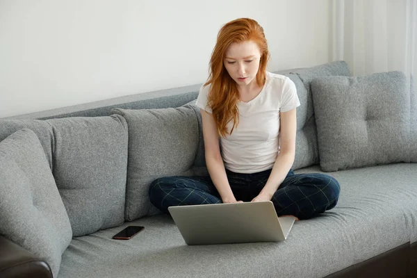 En rödhårig tjej sitter i soffan i hemkläder och tittar in i en laptop.. Stockfoto