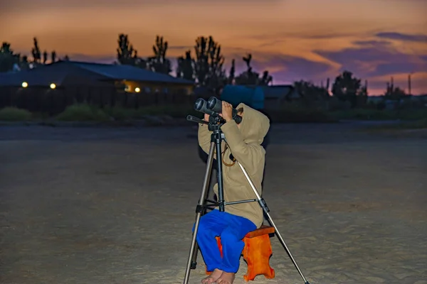 Um menino olha através de binóculos em um tripé contra o fundo de um pôr do sol da noite . — Fotografia de Stock