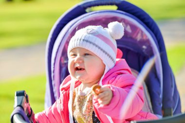 Kırmızı ceketli, beyaz şapkalı küçük neşeli bir kız sonbahar parkında bebek arabasına biniyor..