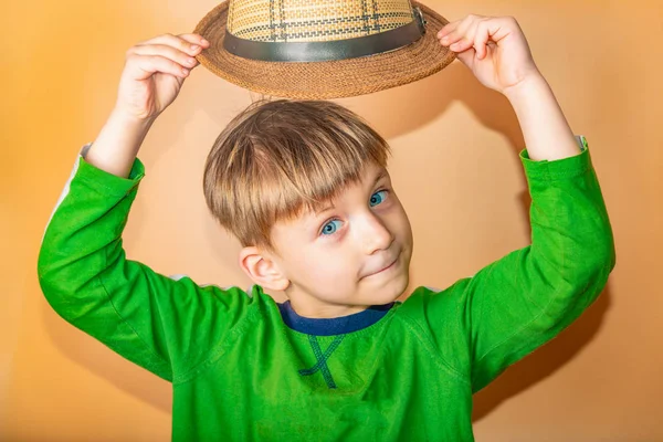 Un niño con un sombrero de paja y ropa verde sobre un fondo beige se quita el sombrero en saludo . —  Fotos de Stock