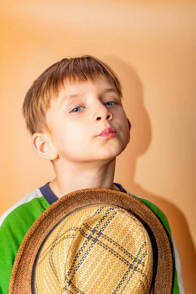 Un niño con un sombrero de paja y ropa verde sobre un fondo beige se quita el sombrero en saludo . —  Fotos de Stock