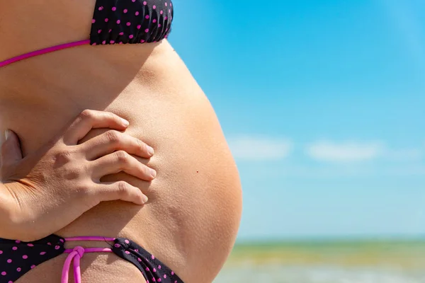 Těhotná dívka drží velké břicho s rukama na pláži u moře. — Stock fotografie
