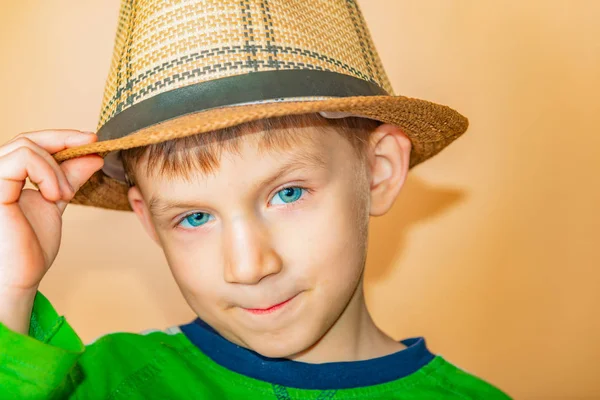 Un niño con un sombrero de paja y ropa verde sobre un fondo beige se quita el sombrero en saludo . —  Fotos de Stock