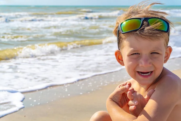 Radosny i szczęśliwy chłopiec w okularach przeciwsłonecznych siedzi na piasku morskim na plaży, uśmiecha się i rozgląda.. — Zdjęcie stockowe