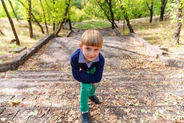 Un niño está de pie en los viejos escalones del parque, apoyado en su rodilla y mirando a la cámara, foto de gran angular . —  Fotos de Stock