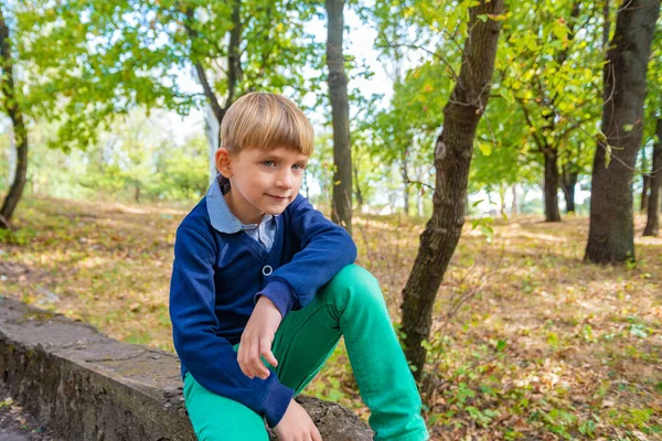 Młody i słodki chłopiec w zielonych dżinsach siedzi na krawężniku w parku z ręką na kolanie. — Zdjęcie stockowe