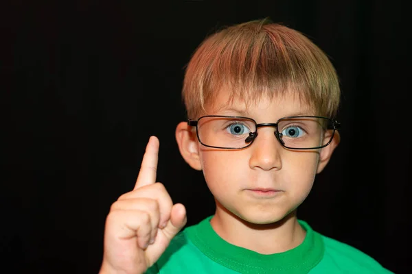 Der Junge mit Brille auf schwarzem Hintergrund blickt in die Kamera und zeigt einen Finger nach oben. — Stockfoto