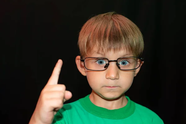 Der Junge mit Brille auf schwarzem Hintergrund blickt in die Kamera und zeigt einen Finger nach oben. — Stockfoto