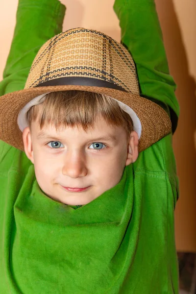 Um menino com um chapéu de palha e roupas verdes em um fundo bege tira o chapéu em saudação . — Fotografia de Stock