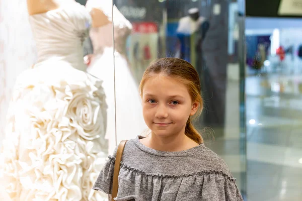 Ein junges Mädchen, ein Teenager, steht am Fenster eines Hochzeitssalons und betrachtet Brautkleider, stellt das Familienleben vor. — Stockfoto