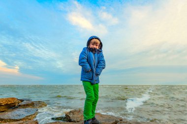 Ceketli donmuş bir çocuk deniz kıyısındaki taşların üzerinde duruyor, geniş açılı fotoğraf..