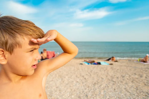 The boy looks into the distance, covering his face with his hand from the sun on the beach, by the sea. — ストック写真
