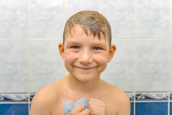 Радостный мальчик после купания в душе с мокрой головой улыбается и держит в руках мочалку — стоковое фото