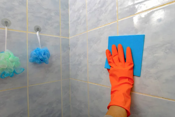 Женщина вытирает плитку синей салфеткой в ванной комнате в оранжевых защитных перчатках . — стоковое фото