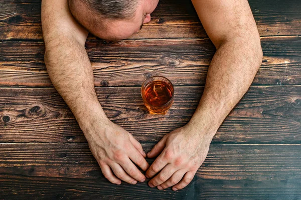 П'яний чоловік спить на дерев'яному столі після сп'яніння, поруч склянка коньяку, віскі, бренді. Поняття алкоголізму та алкогольної залежності . — стокове фото