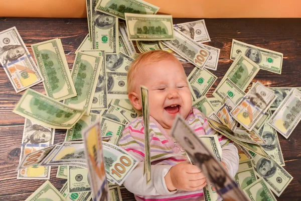 A small child lies on dollar bills, looks at the camera and laughs. The concept of wealth and financial independence.