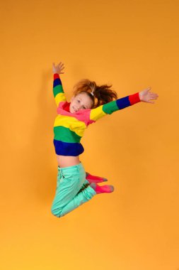 A girl in colored and bright clothes in a jump holds her hands to the sides and tightens her legs on a yellow background.