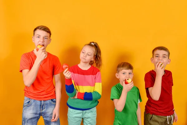 Quattro bambini mangiano una mela in piedi uno accanto all'altro, ragazzi su uno sfondo giallo in abiti colorati . — Foto Stock