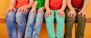 Four children in bright and colorful clothes are sitting with their legs dangling and hands on their knees, on a yellow background.