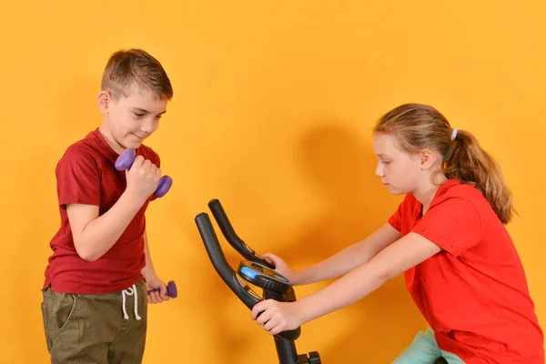 Dambılı bir çocuk ve egzersiz bisikletinde oturan bir kız, sarı arka planda spor yapan pozitif çocuklar.. — Stok fotoğraf