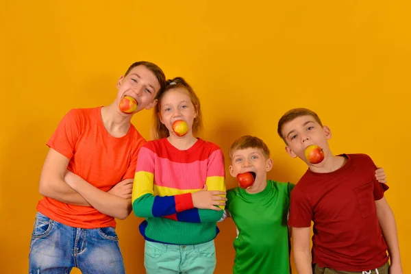 Quatro crianças com uma maçã na boca, irmãos e irmãs em roupas brilhantes e coloridas anunciam um estilo de vida saudável e uma dieta saudável. . — Fotografia de Stock
