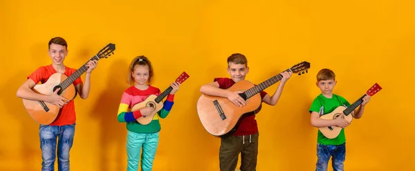 Cuatro niños con guitarras acústicas en ropa brillante y colorida de pie en una fila sobre un fondo amarillo . —  Fotos de Stock