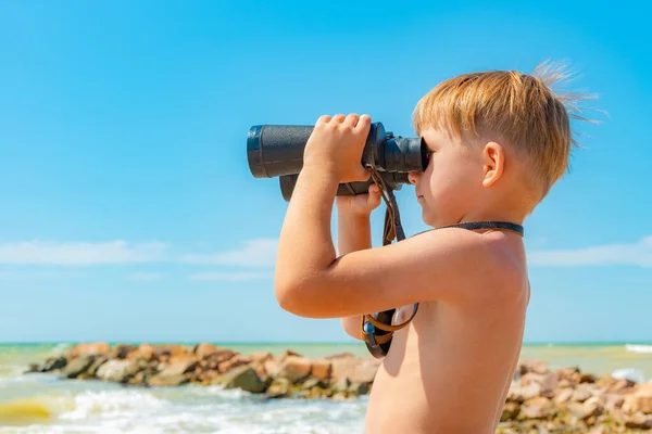 A boy with binoculars looks at the sea against a blue sky.