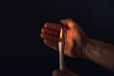 Adamın eli yanan bir mumu kapatır, siyah bir arka planda..