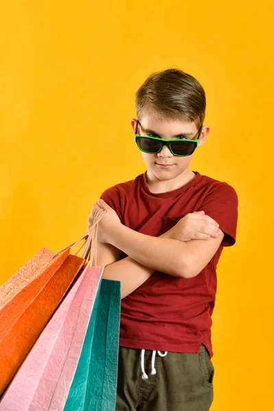 Хлопчик Сонцезахисних Окулярах Кольоровими Мішками Руці Дитина Покупками Жовтому Тлі — стокове фото