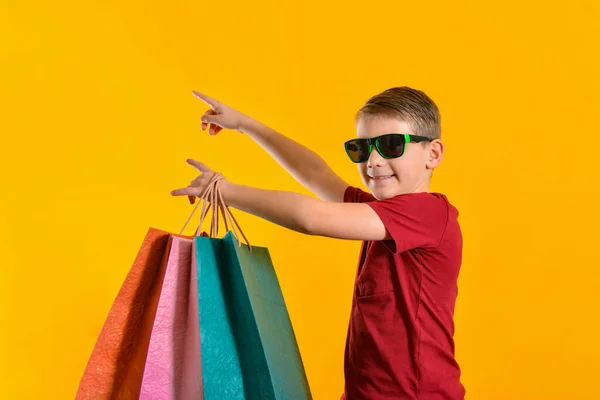 Хлопчик Сонцезахисних Окулярах Кольоровими Мішками Руці Дитина Покупками Жовтому Тлі — стокове фото