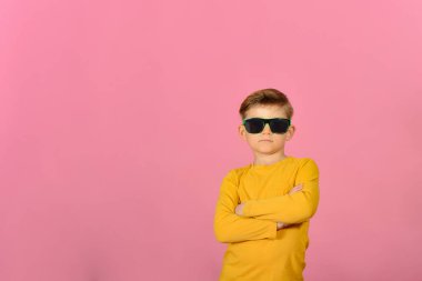 Kameraya poz veren göz kamaştırıcı ve şık bir çocuk. Güneş gözlüklü, sarı elbiseli, pembe arka planda bir model..