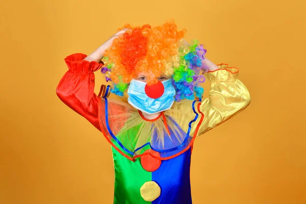 Sad child clown in a protective medical mask against coronavirus on a yellow background holds his hands behind his head and does not know what to do next.