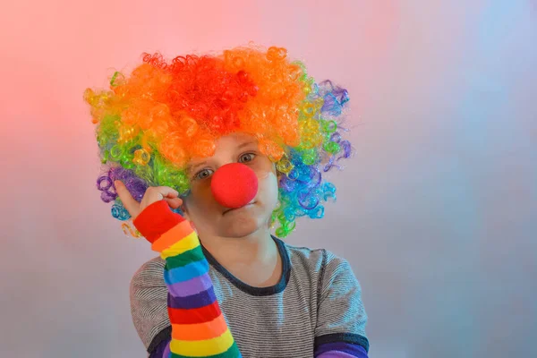 Joyful boy in a clown wig and with a red nose holds his index finger to the top