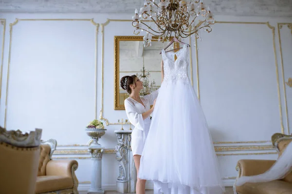 Όμορφη νύφη σε ρόμπα είναι βλέποντας ένα γαμήλιο φόρεμα — Φωτογραφία Αρχείου