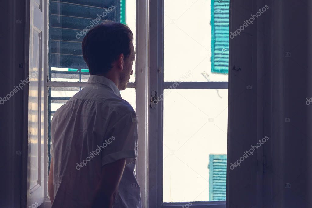 Man looks out window from a dark room 