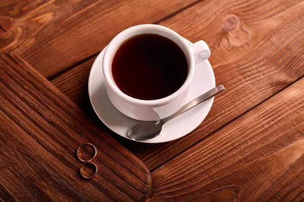 Preto, sabor, café orgânico em copo clássico branco em uma mesa de madeira com um par de alianças de casamento . — Fotografia de Stock