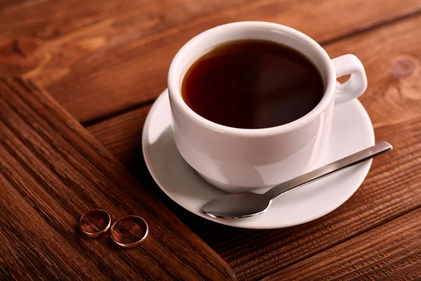 Preto, sabor, café orgânico em copo clássico branco em uma mesa de madeira com um par de alianças de casamento . — Fotografia de Stock