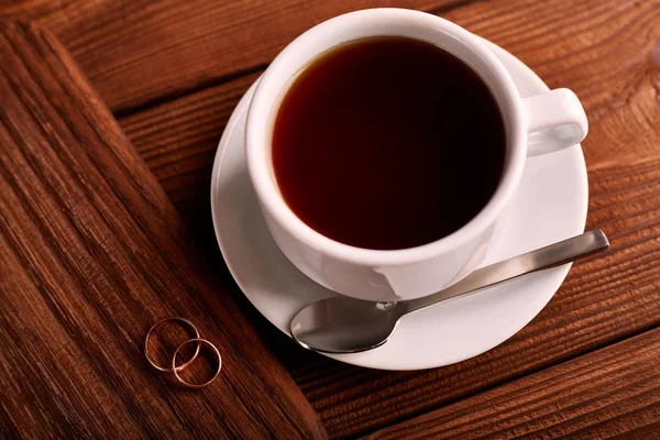 Preto, sabor, café orgânico em copo clássico branco em uma mesa de madeira com um par de alianças de casamento . — Fotografia de Stock